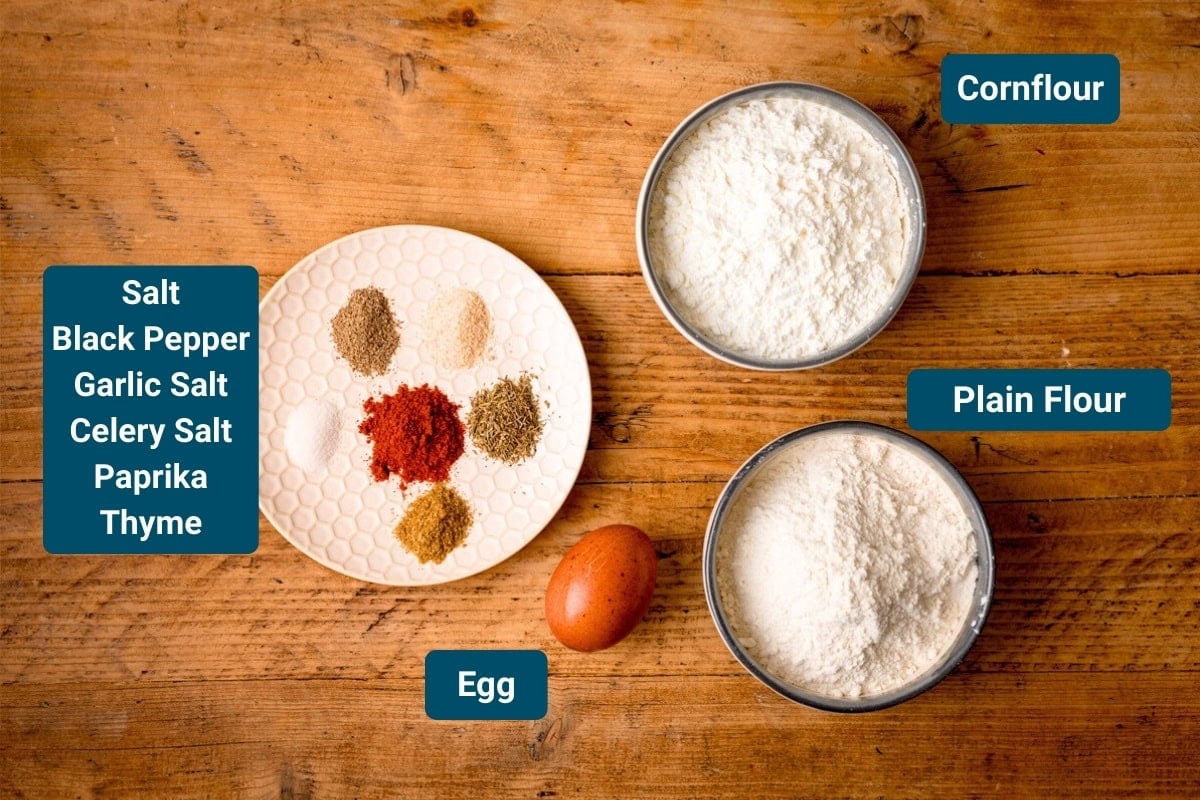 Ingredients for the coating of air fryer popcorn chicken on a wooden surface. The ingredients are labelled and include a bowl of flour, a bowl of cornflour, a plate with spices, and a whole egg.