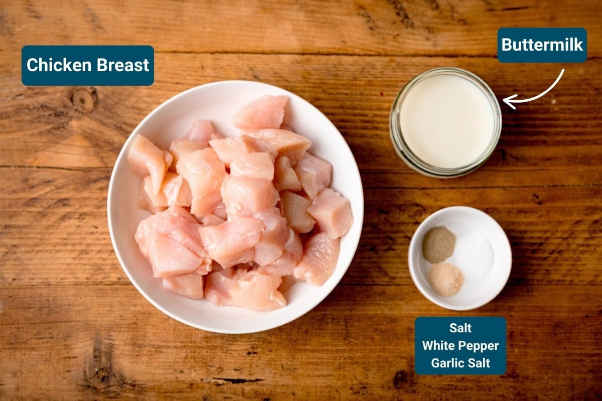 A bowl of raw diced chicken breast, beside a jar of buttermilk and a small dish with salt, white pepper, and garlic salt on a wooden surface. Ingredients are labelled.