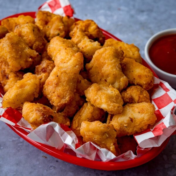 A red basket lined with paper holds a pile of crispy air fryer popcorn chicken next to a small bowl of tomato ketchup.
