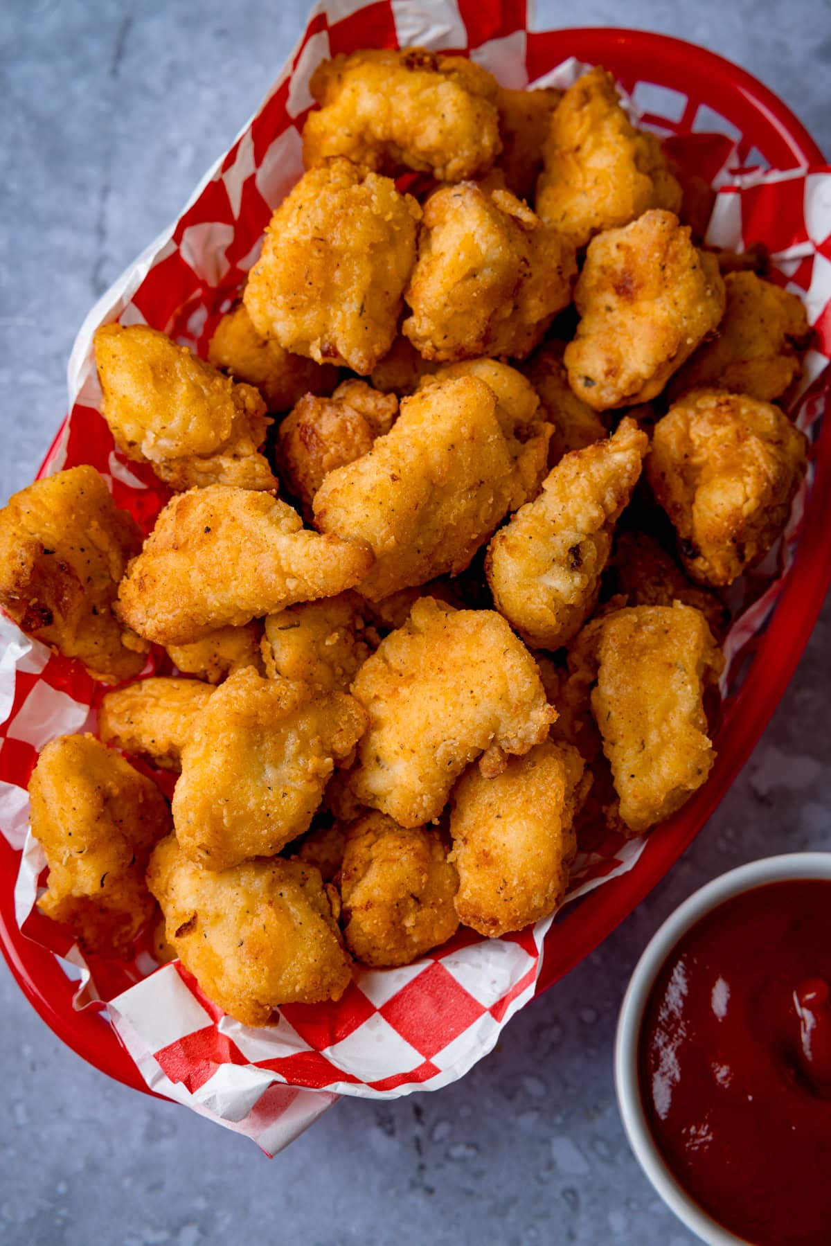 A red basket lined with red and white chequered paper filled with crispy air fryer popcorn chicken nuggets, next to a small bowl of tomato sauce.
