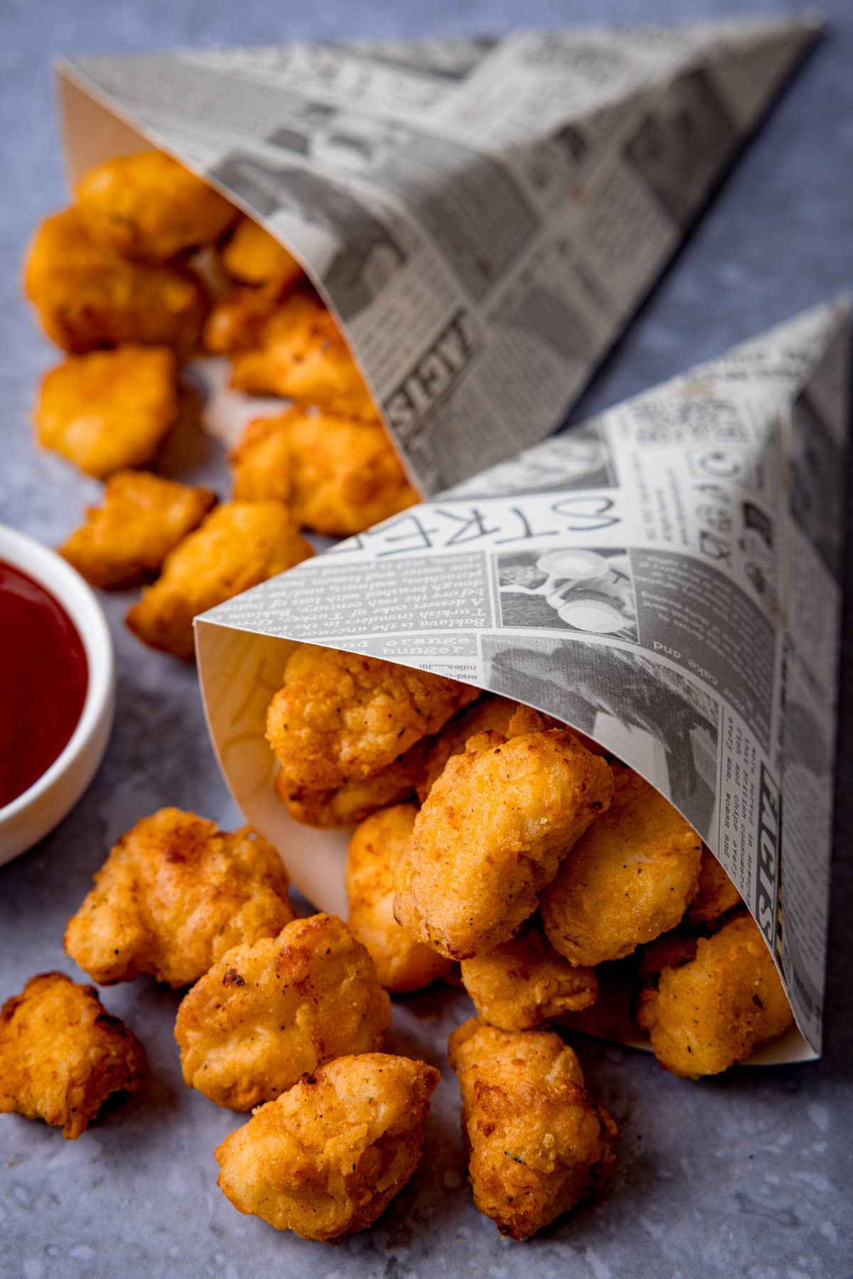 Two paper cones filled with crispy Popcorn Chicken spilling onto a grey surface, accompanied by a small bowl of ketchup.