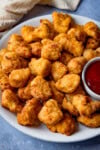 A plate of golden-brown popcorn chicken with a small bowl of ketchup on the side on a light blue surface.