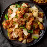 A plate of Salt and Pepper Chicken pieces with onions, chillies, and spring onions on a dark plate.