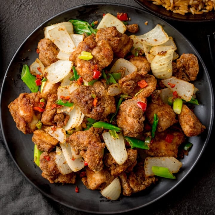 A plate of Salt and Pepper Chicken pieces with onions, chillies, and spring onions on a dark plate.