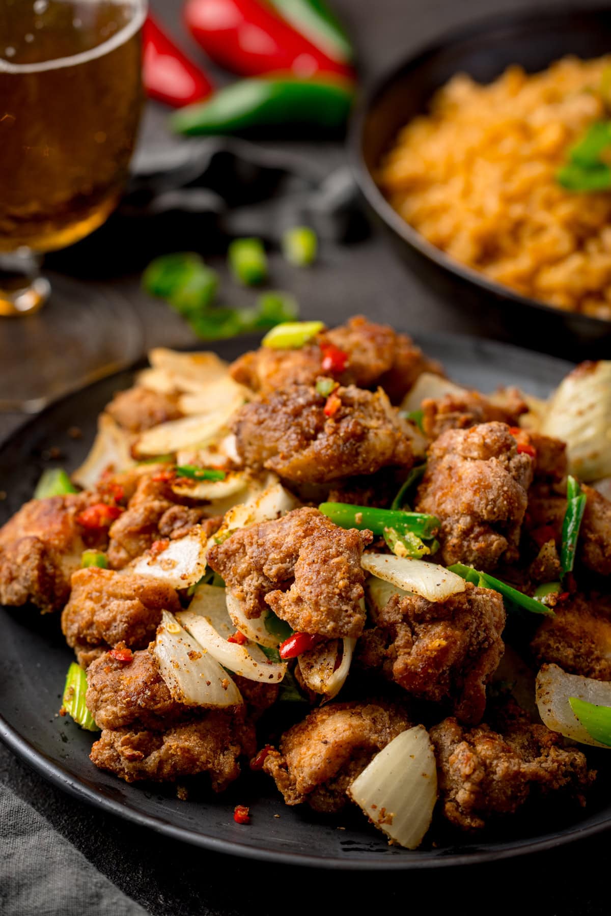 Tall image of a plate of crispy Salt and Pepper Chicken pieces with onions, spring onions, and red chillies on a dark background. There is a plate of rice, a glass of beer and some fresh chillies partially visible in the background.