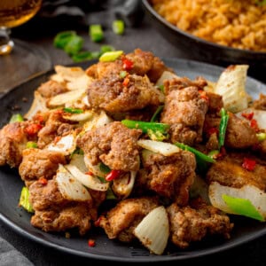 Wide image of a plate of crispy Salt and Pepper Chicken pieces with onions, spring onions, and red chillies on a dark background. There is a plate of rice partially visible in the background.
