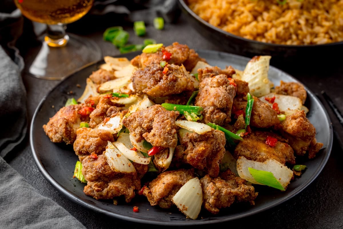 A black plate with chunks of crispy chicken, onions red and green chilies and scallions.