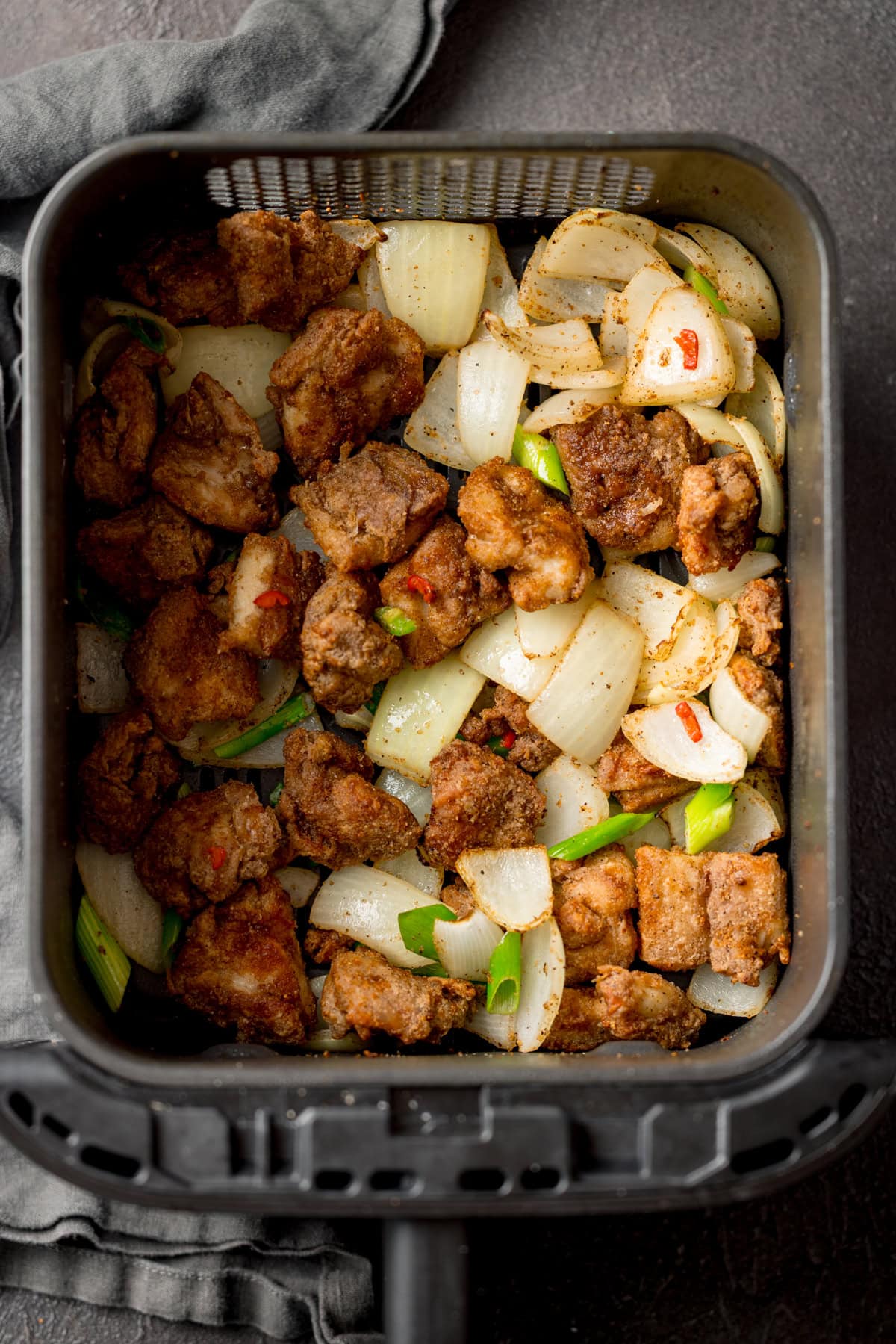 Salt and Pepper Chicken pieces with chopped onion, scallions and chillies in an air fryer basket, seen from above.