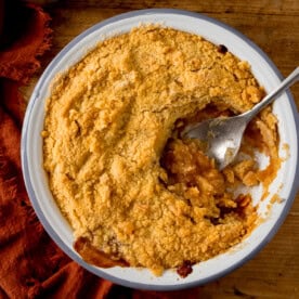 A round baking dish filled with golden-brown apple crumble. A scoop is being taken by a silver serving spoon. The dish is on a wooden surface next to a terracotta napkin.