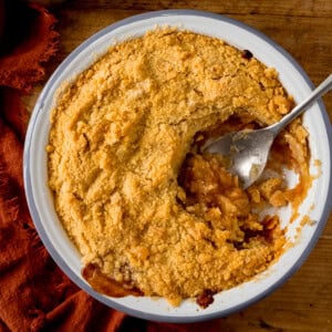 A round baking dish filled with golden-brown apple crumble. A scoop is being taken by a silver serving spoon. The dish is on a wooden surface next to a terracotta napkin.
