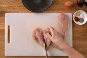 A hand slices raw chicken breast fillets on a white chopping board; an egg, spices, and a bowl sit on the wooden worktop nearby.