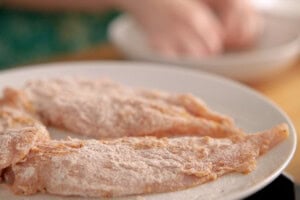 Raw chicken strips coated in seasoned flour are arranged on a white plate, with a person's hands preparing food in the blurred background.