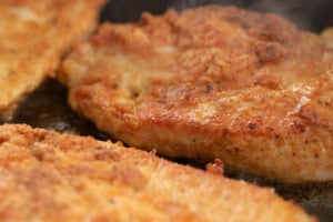 Close-up of seasoned chicken breast fillets cooking in a frying pan with visible browning and spices on the surface.