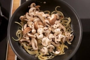 Sliced mushrooms and onions cooking in a black frying pan on a hob.