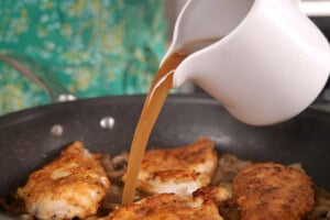 A person pours chicken stock from a white jug into a pan with browned chicken pieces.