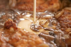 Close-up of a cream being poured over browned pieces of chicken, onions, and mushrooms in a frying pan.
