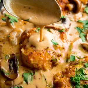 A spoon pours creamy Chicken Marsala sauce over browned chicken fillets, garnished with chopped parsley.