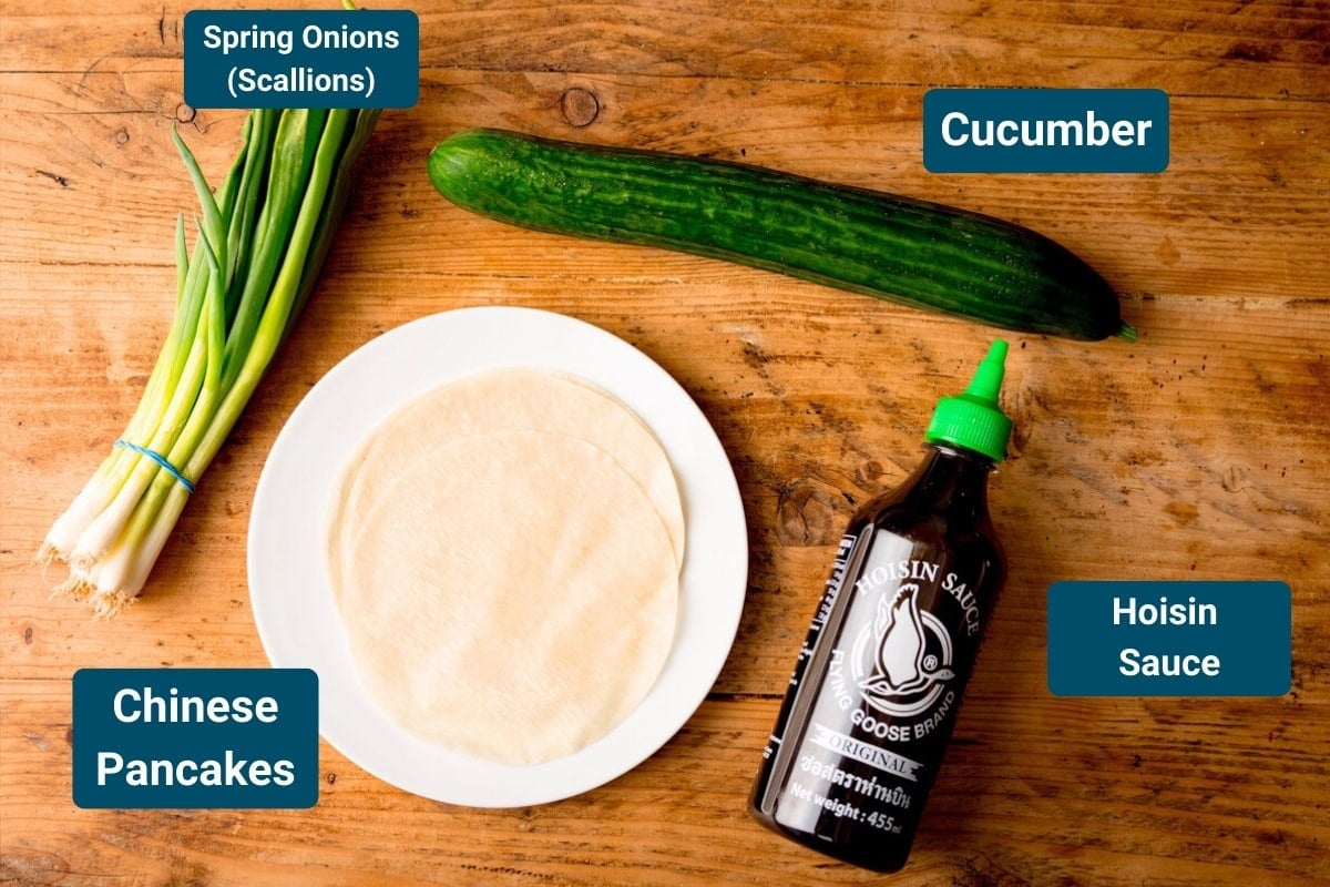 A wooden table with spring onions, a cucumber, a plate of Chinese pancakes for crispy duck, and a bottle of hoisin sauce, each labelled with text.