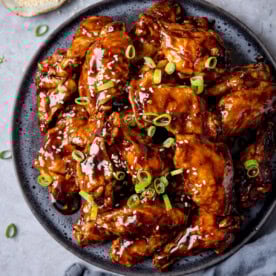A plate of tamarind chicken wings glazed to perfection, garnished with sliced spring onions and sesame seeds, served on a dark round dish.