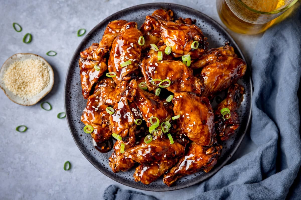 A plate of Tamarind Chicken Wings glazed to perfection, garnished with chopped spring onions and sesame seeds, sits next to a small bowl of sesame seeds and a refreshing drink.