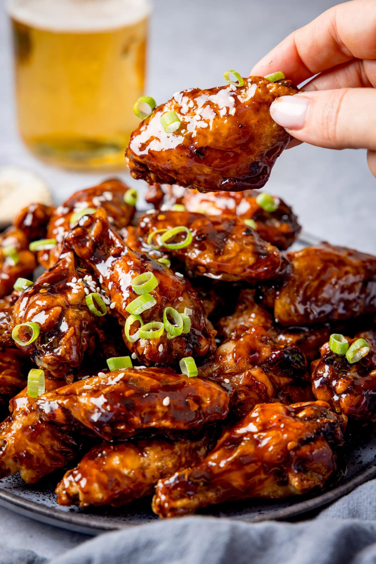 A plate of tamarind chicken wings glazed to perfection and topped with chopped spring onions, a hand picking up one wing; a glass of beer in the background.