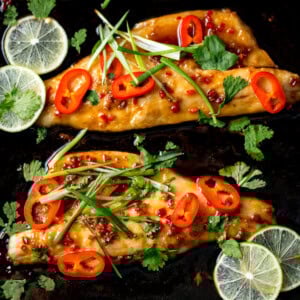 Two fillets of Asian Sea Bass, glazed and baked, topped with sliced red chilli, spring onions, and coriander, garnished with fresh lime slices on a dark background.