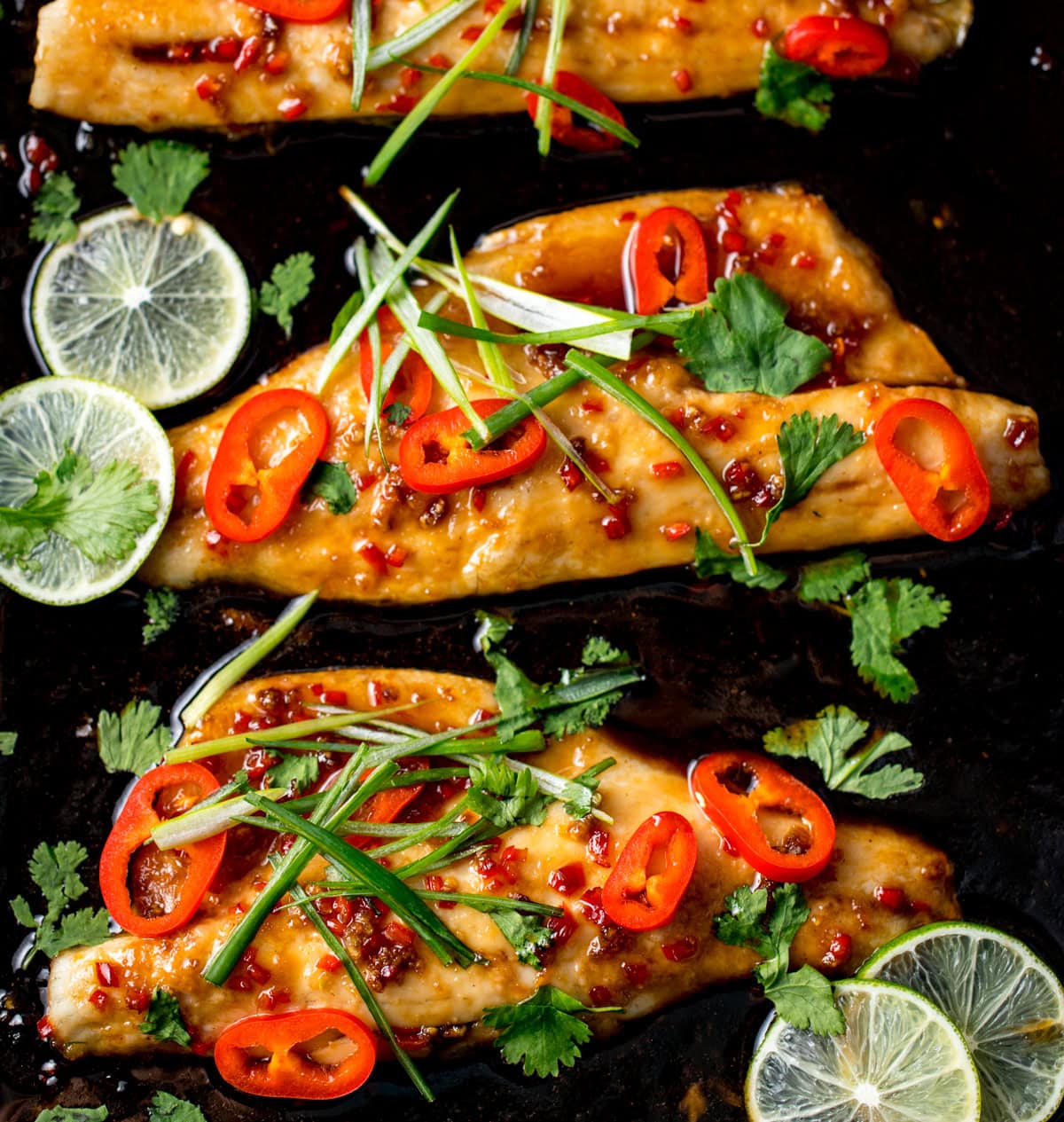 Baked sea bass fillets topped with sliced red chilli, spring onions, and coriander, garnished with lime slices on a dark baking tray.