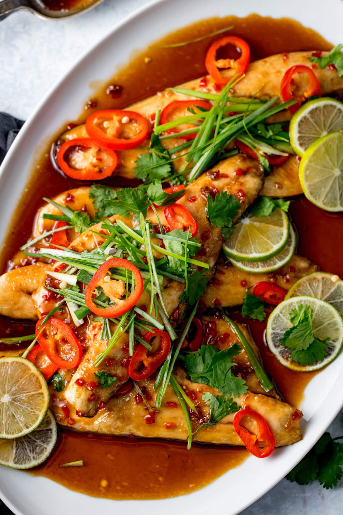 Oven-baked sea bass fillets topped with sliced red chilli, lime, coriander, and spring onions, served with a sticky glaze on a white platter.