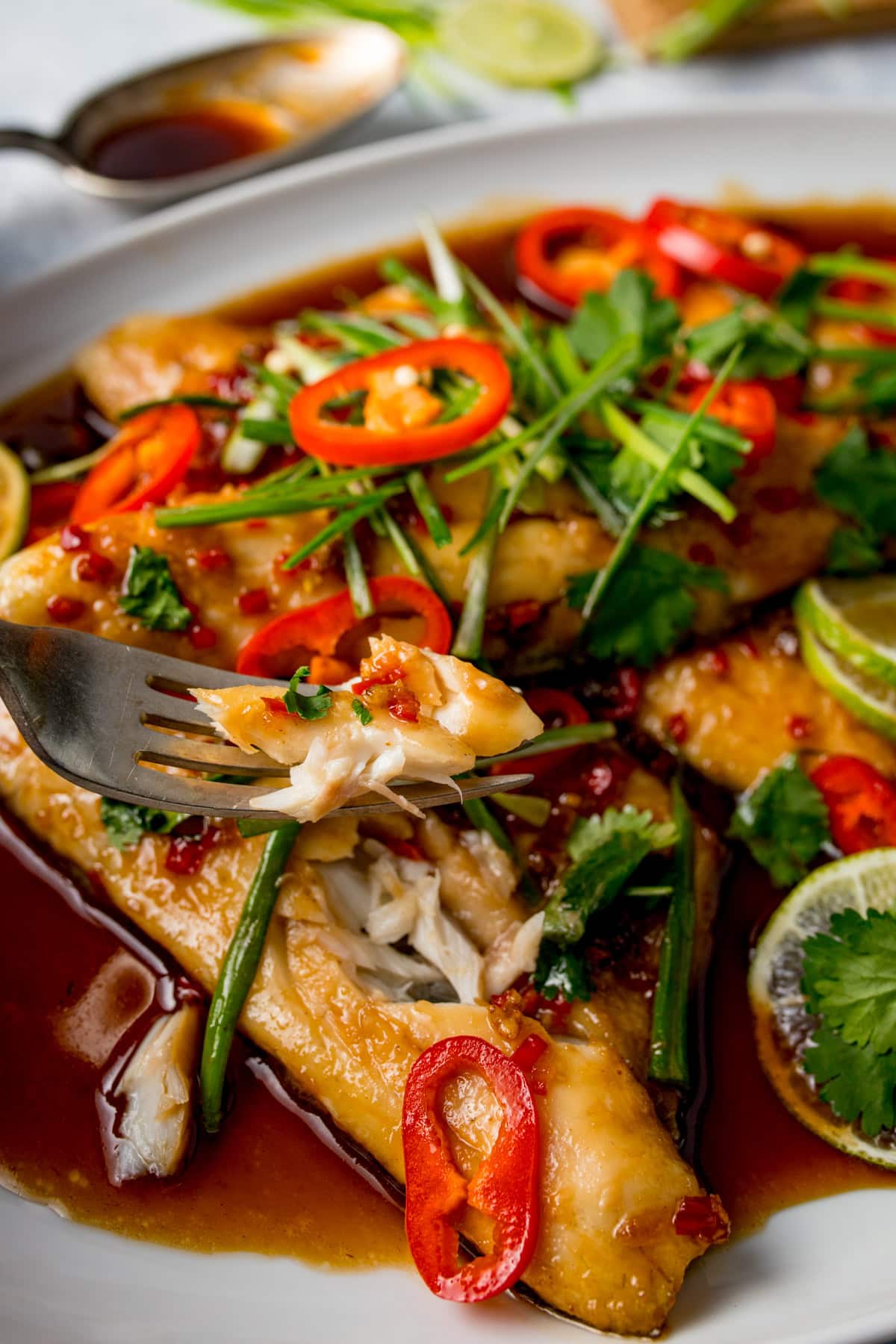 A close-up of Chinese Sea Bass fillets, oven baked and topped with sliced red chilli, herbs, and spring onions in a soy-based sauce, with a fork lifting a flaky piece.
