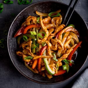 Overhead image of a black bowl filled with Black Pepper Chicken, Chinese-style stir-fried with sliced red and green peppers, onions, and spring onions, served with chopsticks.