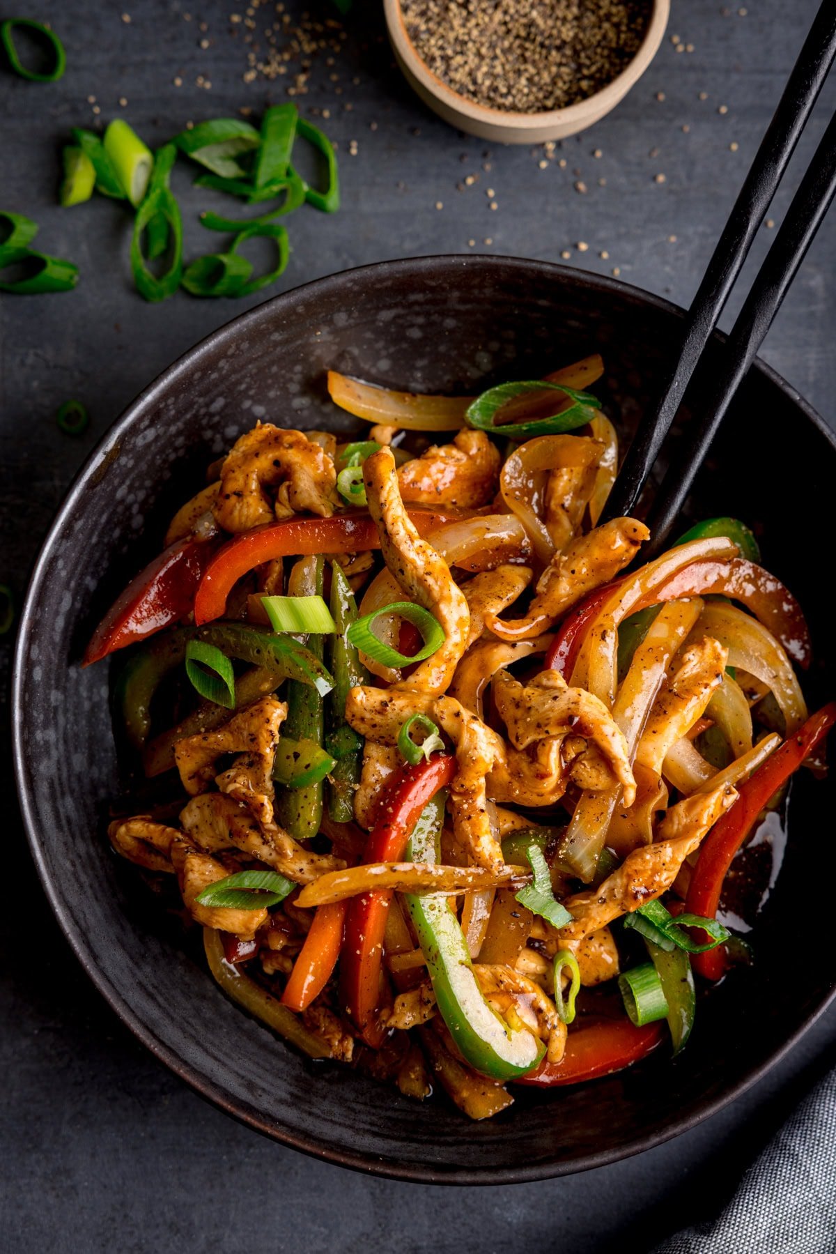 A black bowl on a dark background filled with black pepper chicken, peppers, onions, and spring onions, served with black chopsticks. There are scattered spring onions (scallions) and a little bowl of ground black pepper at the top of the frame.