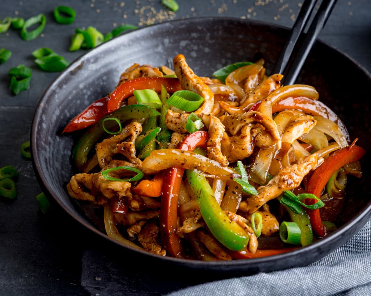 A black bowl filled with chicken in black pepper sauce stir-fried with sliced red and green peppers, onions, and chopped spring onions, served with black chopsticks.