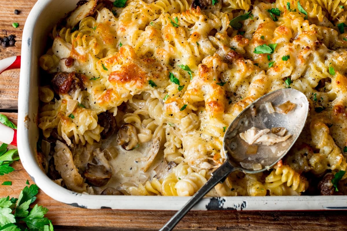 Baked creamy chicken pasta bake with spirali, mushrooms, cheese, and herbs in a white dish, served with a metal spoon on a wooden surface.