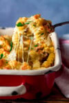 A fork lifting a serving of creamy chicken pasta bake with spiral pasta from a red and white casserole dish.