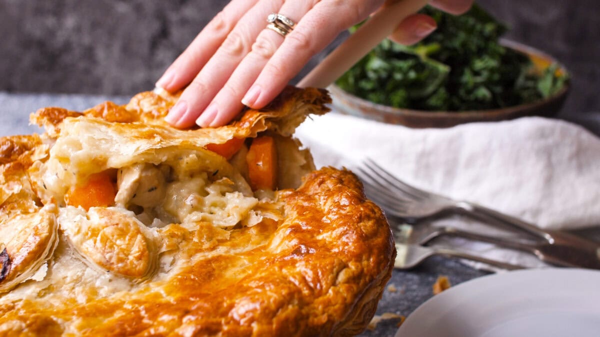 A hand lifts the flaky pastry of a golden-brown creamy chicken pie, revealing its rich filling with chicken and vegetables; a bowl of kale sits in the background.