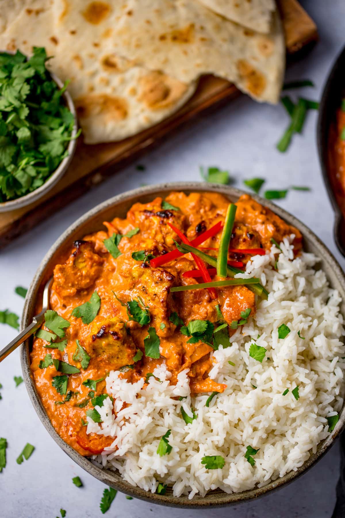 A bowl Chicken Tikka Masala and rice, garnished with coriander and red chilli, served with naan bread and fresh coriander on the side.