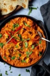 A frying pan of Chicken Tikka Masala, garnished with coriander and red chilli slices, with a serving spoon and naan bread on the side.