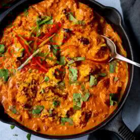 A frying pan filled with Chicken Tikka Masala, garnished with coriander and sliced red chilli, with a spoon resting inside.