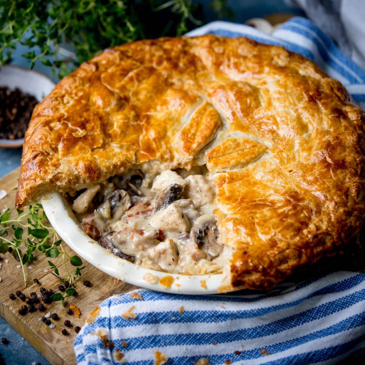 Golden-brown chicken and mushroom pie with a flaky pastry, one slice missing, showing creamy filling; on striped cloth with herbs and peppercorns.