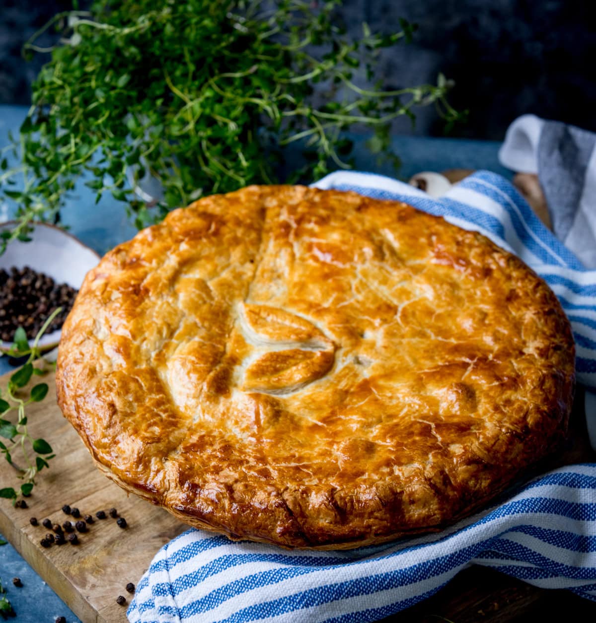 A golden-brown chicken and mushroom pie with a leaf design on top sits on a blue-striped kitchen tea towel, with herbs and peppercorns nearby.