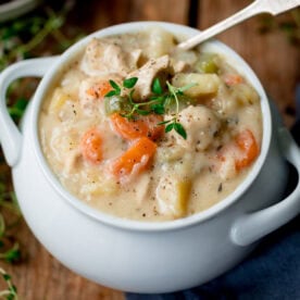 A white bowl filled with creamy chicken pot pie soup containing pieces of chicken, carrots, celery and potatoes, topped with fresh thyme.