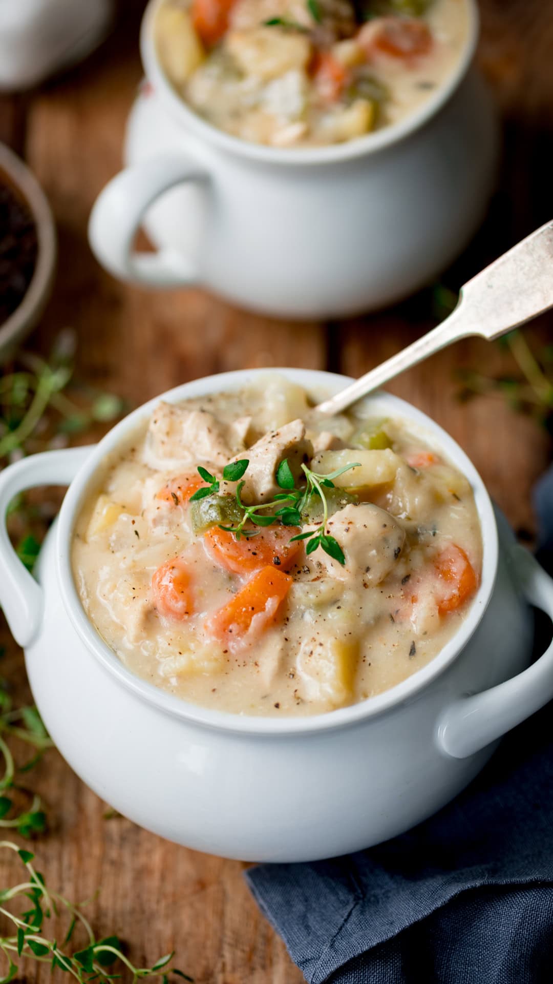 A white bowl filled with creamy chicken pot pie soup, topped with fresh herbs, with a spoon placed inside. Another bowl is blurred in the background.