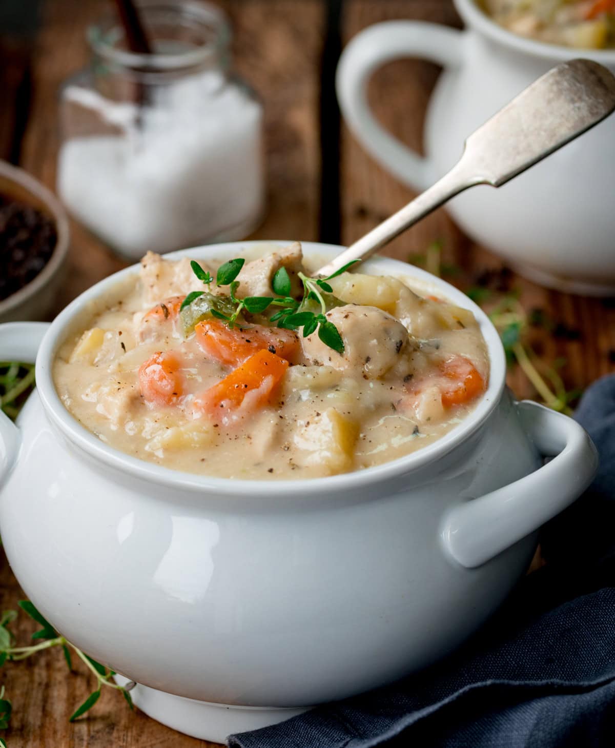 A bowl of creamy chicken pot pie soup with vegetables, garnished with herbs, sits on a wooden table with a spoon in the bowl. A jar of salt and another bowl are in the background.