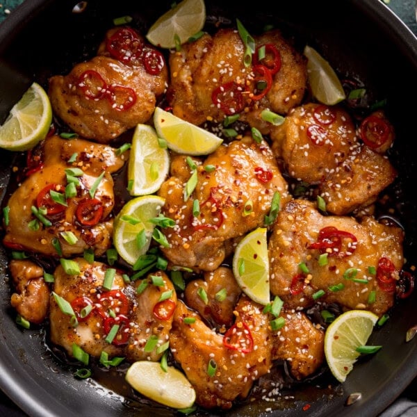 A frying pan filled with chilli lime chicken garnished with sliced red chilli, lime wedges, sesame seeds, and chopped spring onions.