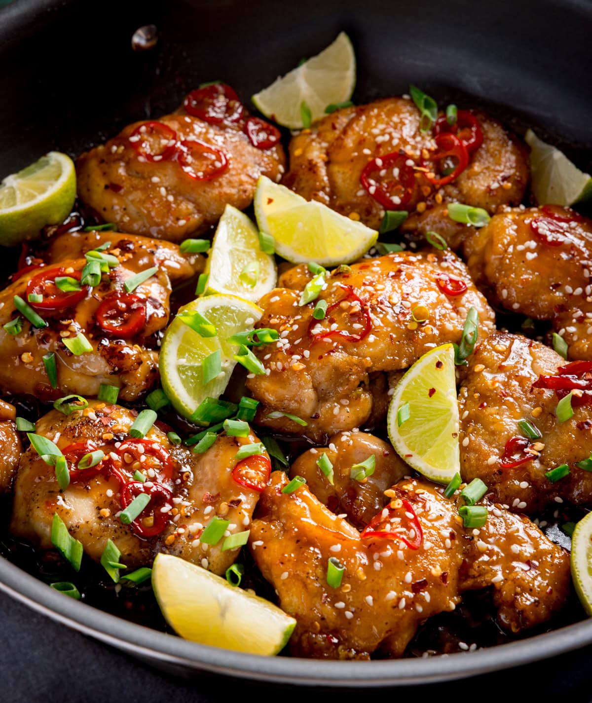 Chilli lime Chicken thighs glazed with sauce, garnished with chopped spring onions, red chilli slices, sesame seeds, and lime wedges in a black frying pan.