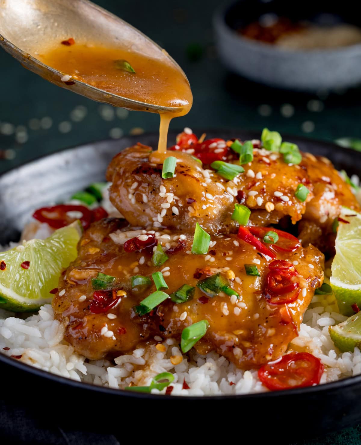 A spoon pours sauce over glazed chilli lime chicken topped with sesame seeds, sliced spring onions, and red chilli, served on white rice with lime wedges.