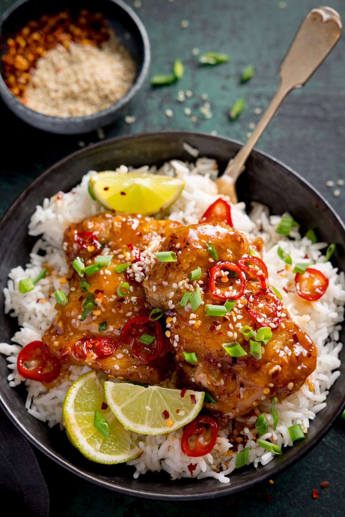 A bowl of white rice topped with glazed chicken, sliced red chilli, spring onions, sesame seeds, and lime wedges, with a fork and a small bowl of seasonings in the background.