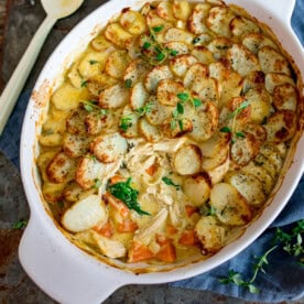 Overhead image of a chicken and potato bake, in a white oval dish, with sliced baked potatoes on a creamy chicken and vegetable filling. A spoonful of the dish has been removed.