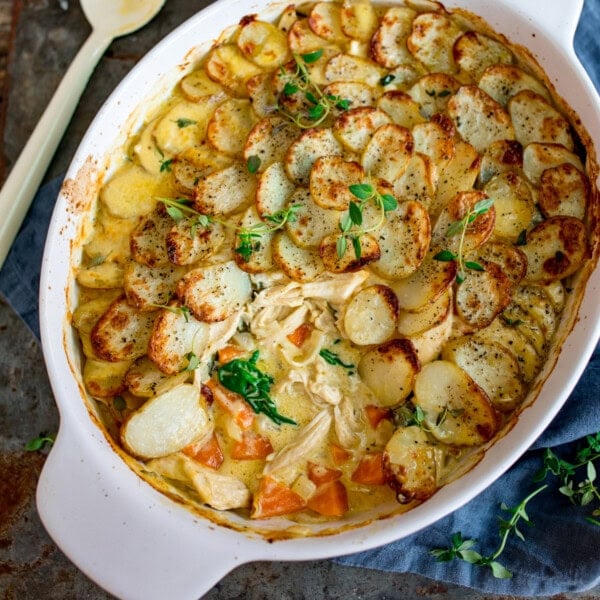 Overhead image of a chicken and potato bake, in a white oval dish, with sliced baked potatoes on a creamy chicken and vegetable filling. A spoonful of the dish has been removed.