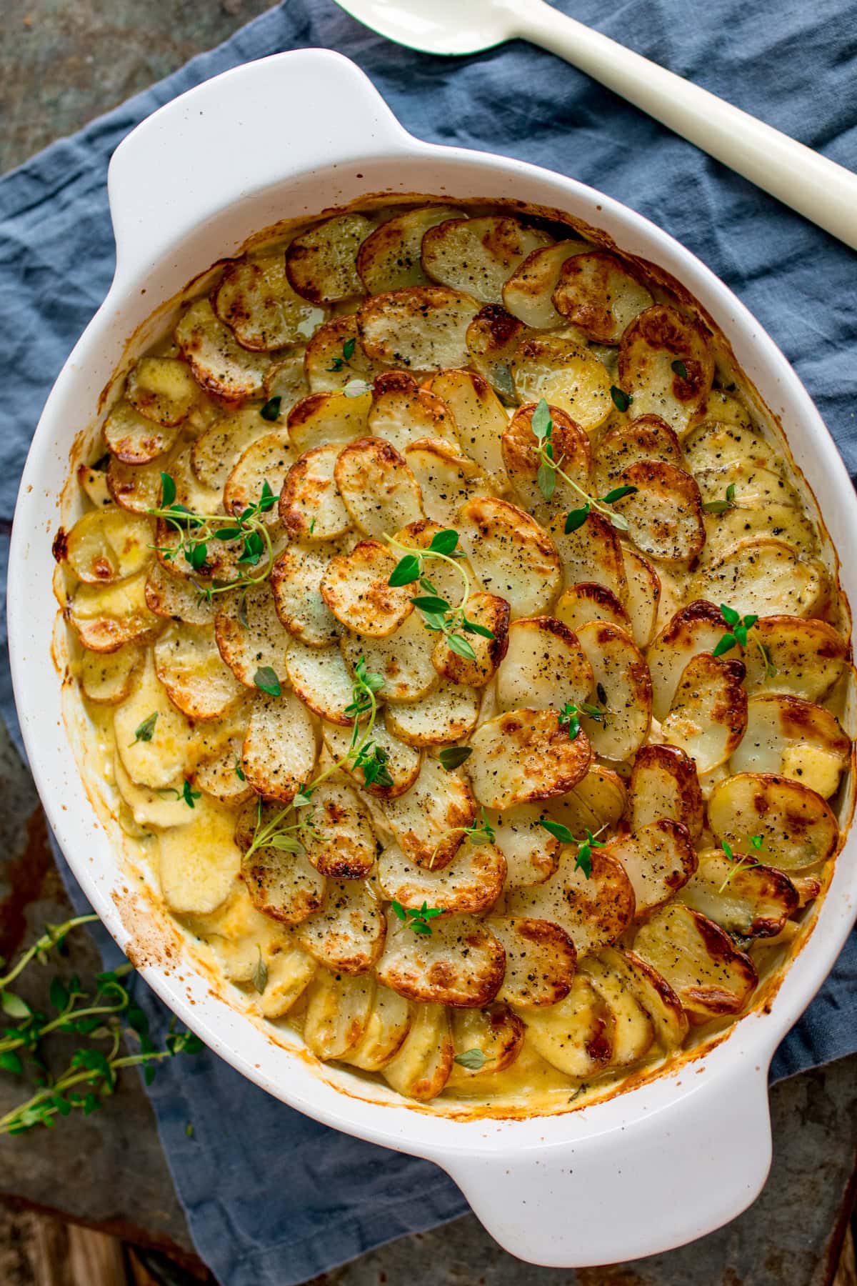 Chicken and Potato Bake served in a white oval dish, topped with sliced, browned potatoes and garnished with fresh herbs. The dish sits on a blue napkin next to a white serving spoon.
